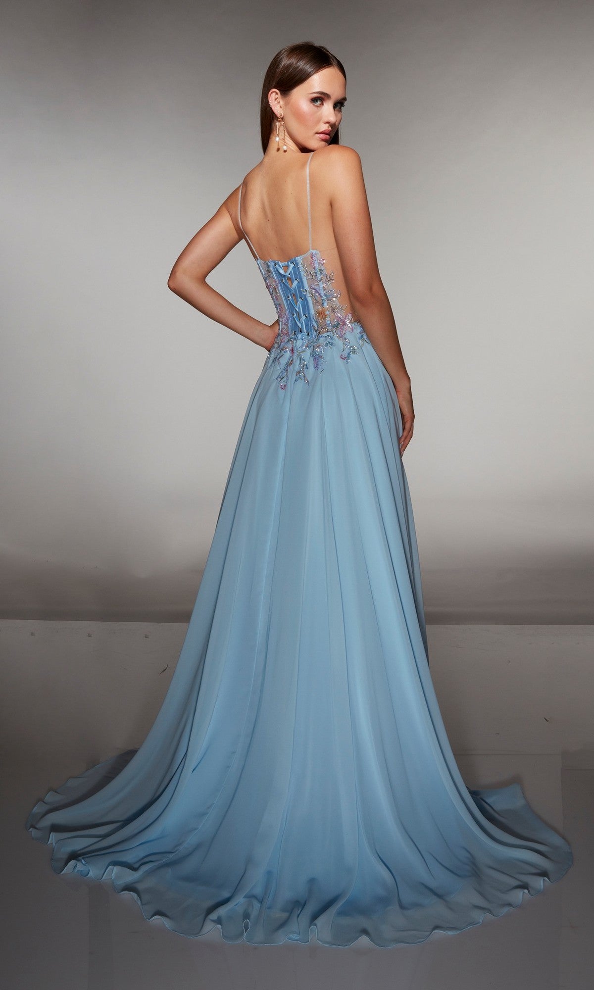 A woman stands in a studio, facing away from the camera, wearing the Alyce 62179 long prom dress in light blue with thin straps and delicate floral appliques on the bodice.